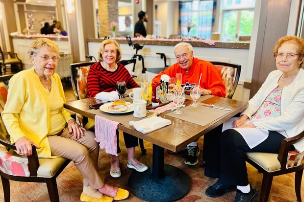 Four residents gather around a table for a festive meal, laughing and enjoying drinks together, reflecting the resort-style senior living Woodbridge hospitality found in this senior community near Prince William Forest Park.