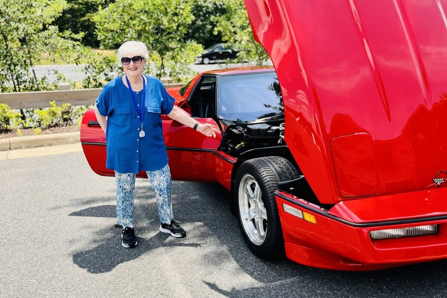 A resident poses smiling beside a classic red sports car with its hood open, enjoying a sunny outdoor car show that reflects the vibrant 55+ senior living Woodbridge lifestyle and convenience of senior living near Potomac Mills.