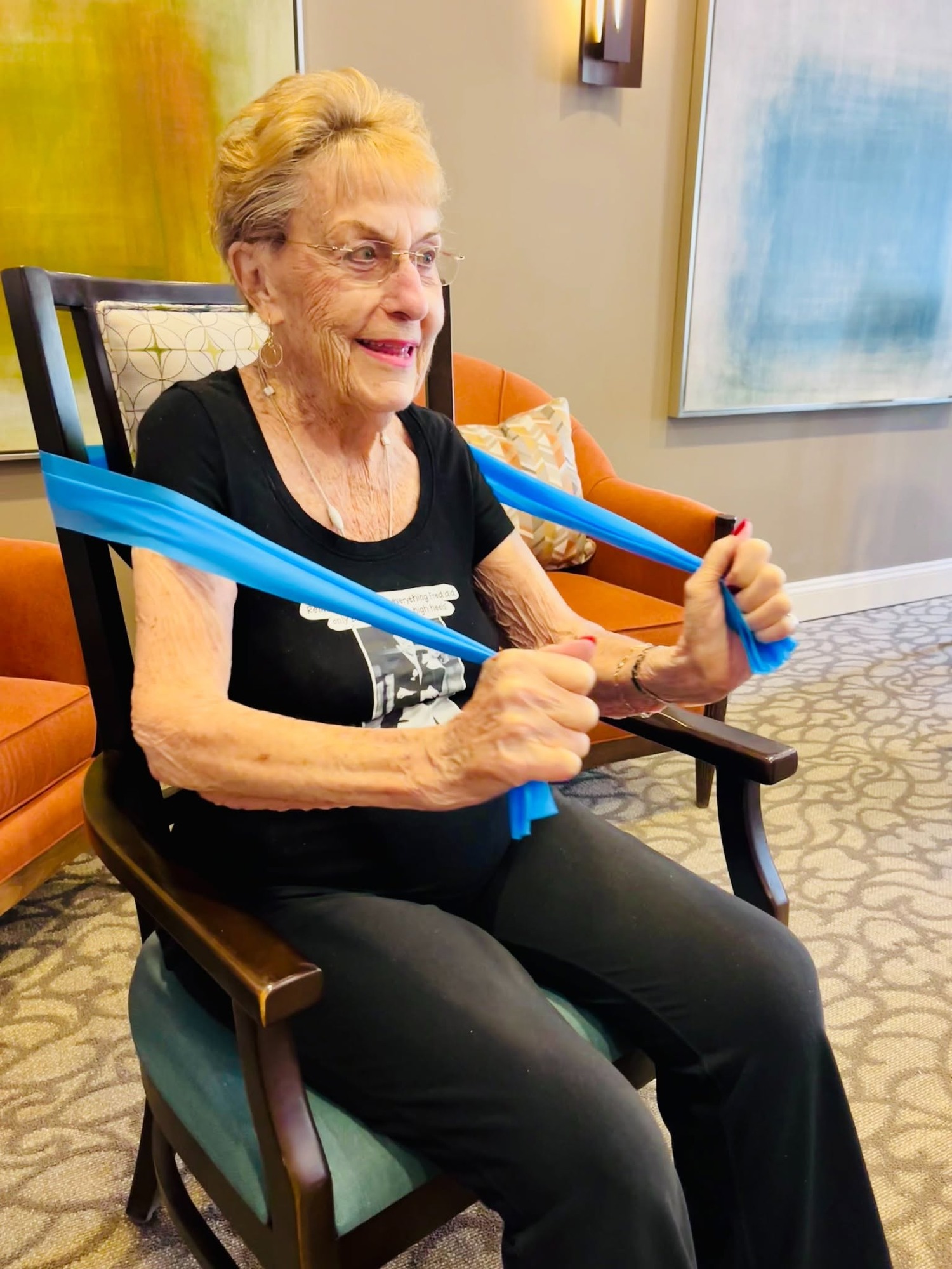 A resident stretches a blue resistance band while seated in a rust-colored armchair, staying active during a seated exercise session that reflects the wellness-focused senior living Northern Virginia programs at this community.