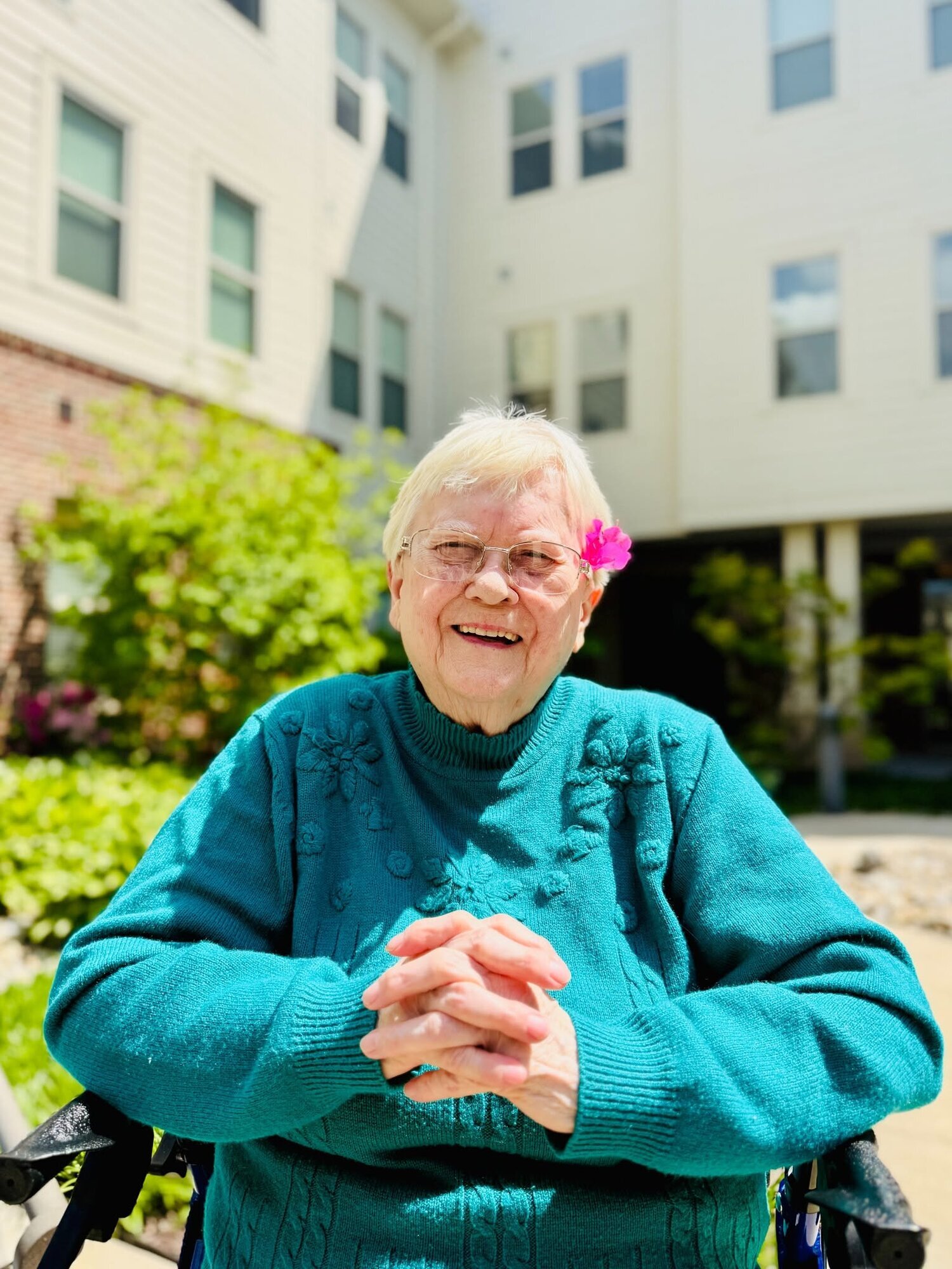 A resident with a pink flower tucked in her hair smiles brightly outdoors, clasping her hands together in front of a sunlit courtyard building, reflecting the warm joy found within this active senior community Prince William County and senior living Woodbridge VA.