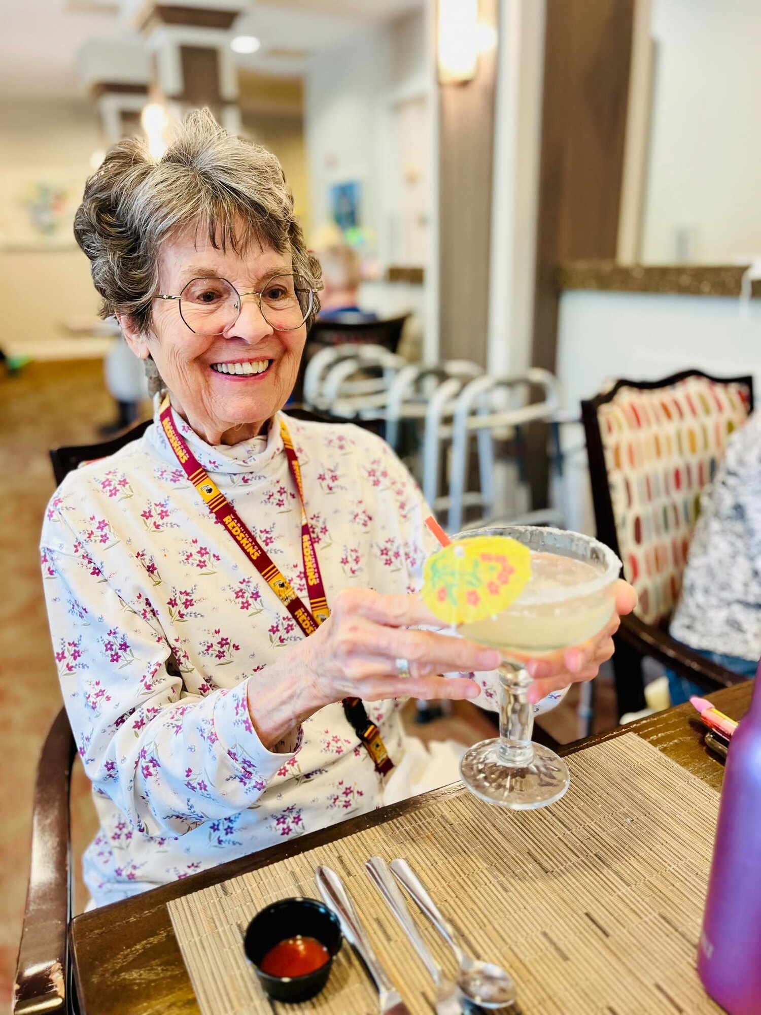 A resident smiles while holding a festive garnished drink at a dining table set with silverware, capturing the resort-style senior living Woodbridge hospitality and convenience of senior living near Potomac Mills.