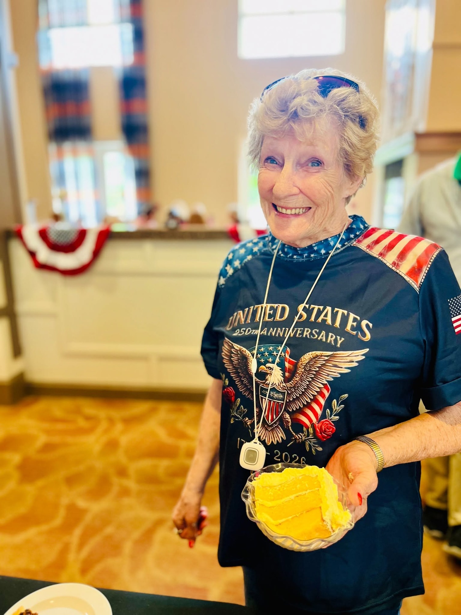A resident smiles while holding a slice of yellow cake, wearing a patriotic red, white, and blue shirt during a festive celebration, reflecting the joyful spirit of senior living Woodbridge VA and this active senior community Prince William County.