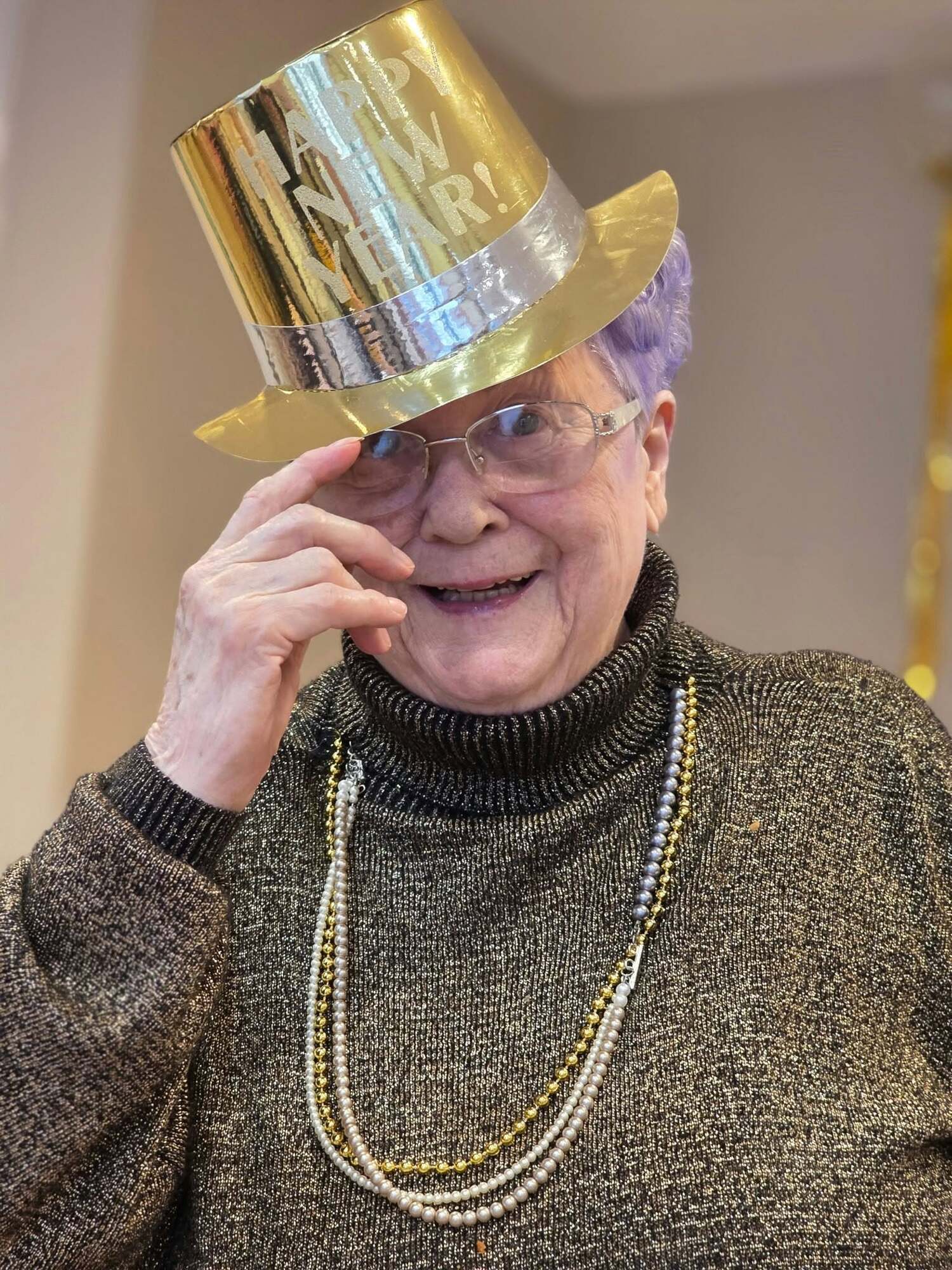 A resident smiles while adjusting a shiny gold New Year's hat, wearing a cozy sweater and pearl necklace, celebrating a festive holiday tradition within this active senior community Prince William County and senior living Woodbridge VA.