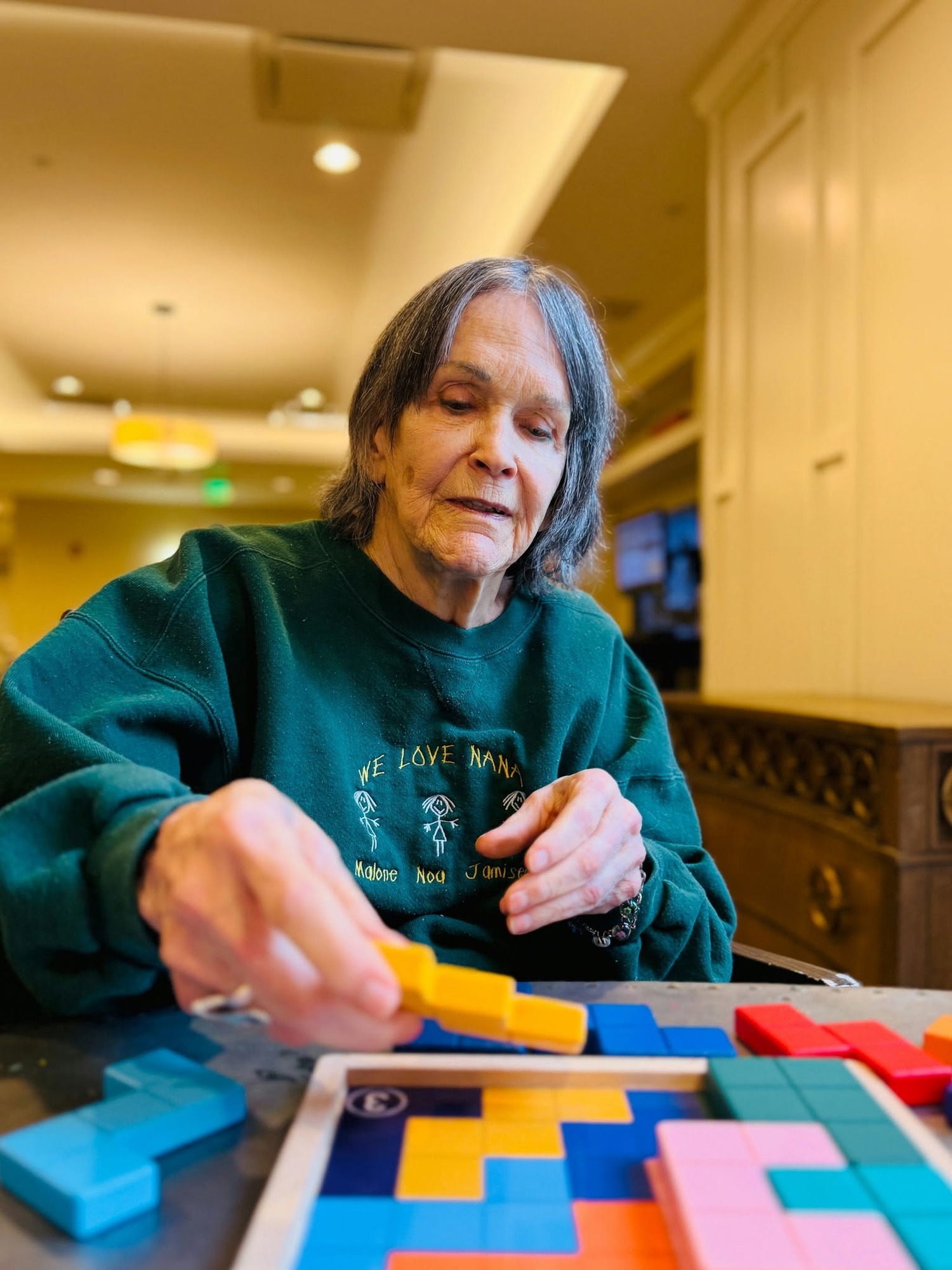A resident wearing a personalized sweatshirt carefully places colorful puzzle pieces onto a board, enjoying a favorite pastime that reflects the vibrant 55+ senior living Woodbridge lifestyle within this active senior community Prince William County.