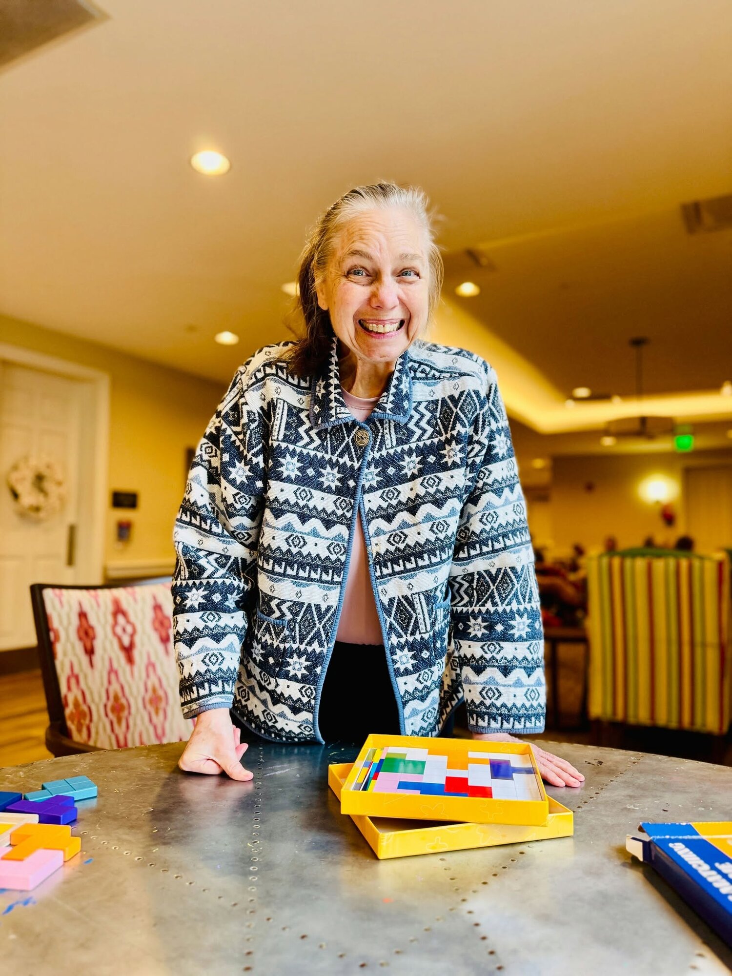 A resident smiles brightly while standing at a table filled with colorful wooden puzzle pieces, enjoying an engaging brain game that reflects the vibrant 55+ senior living Woodbridge lifestyle within this active senior community Prince William County.