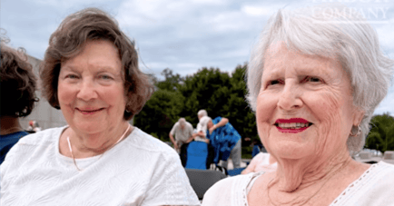 Two women smiling outdoors at an Arbor Company community event, seated with other residents and event tents visible in the background under a partly cloudy sky. The woman on the left has short wavy brown hair and wears a white textured top with a gold necklace. The woman on the right has short white hair, red lipstick, and wears a white lace-trimmed top with a gold brooch and gold hoop earrings.
