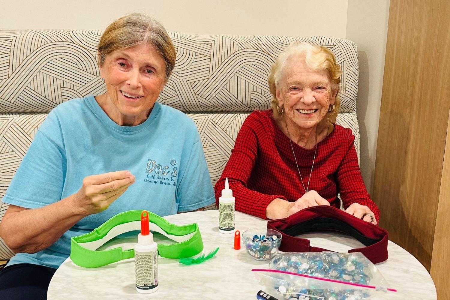 two women smile as they sit at a table and do a craft/make a hat