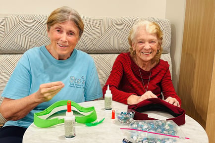 two women smile as they sit at a table and do a craft/make a hat