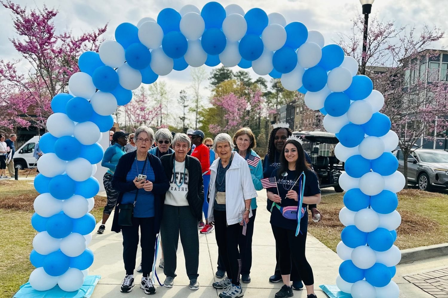 A cheerful group gathers under a blue and white balloon arch on a sunny walkway lined with blooming pink redbud trees, some waving small American flags, a festive outing that showcases senior living with walkable neighborhoods Atlanta, GA and active senior living Atlanta, GA.