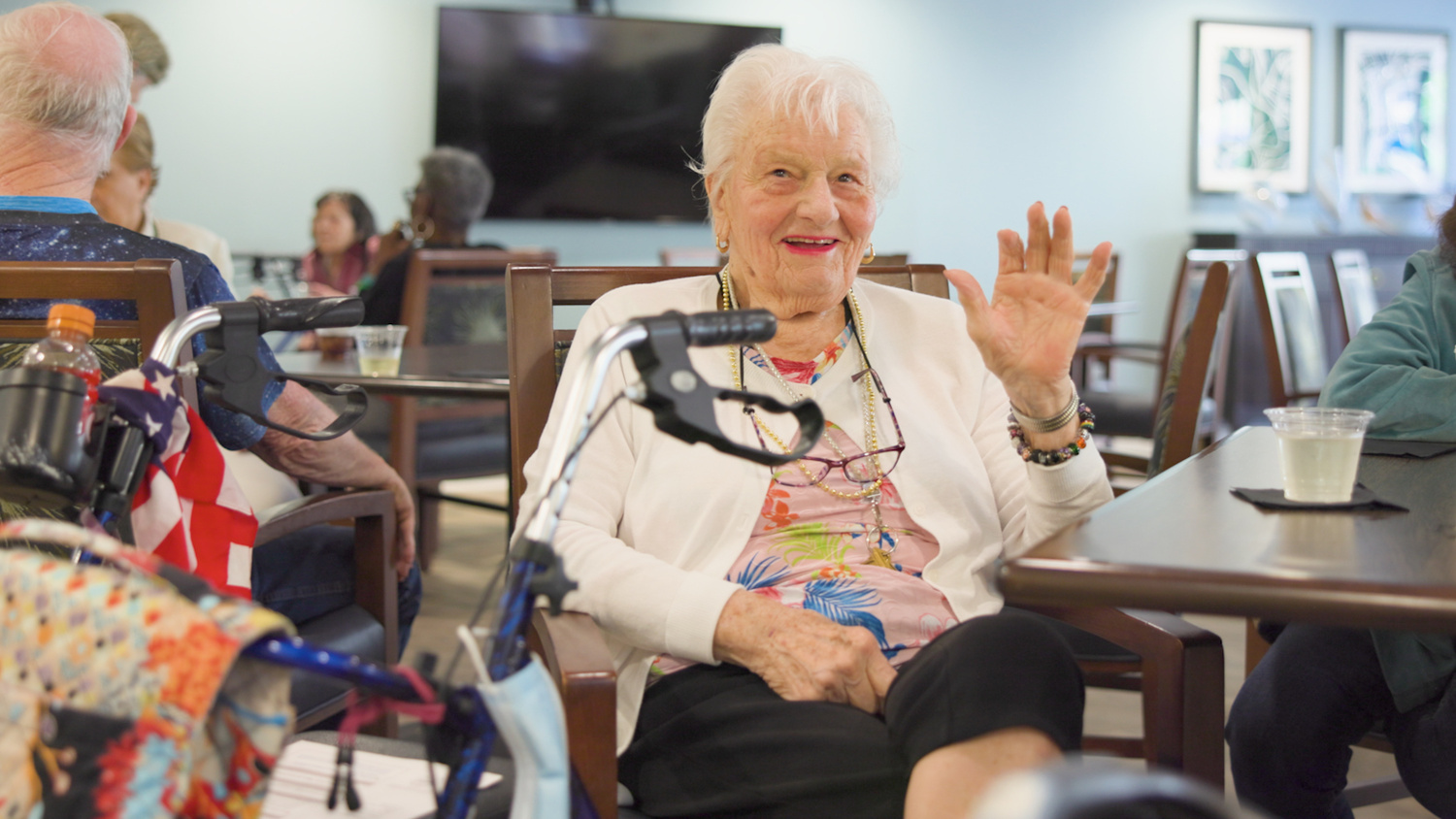 A smiling senior woman waves cheerfully while seated at a dining table in a bright senior living community, enjoying time with others nearby.