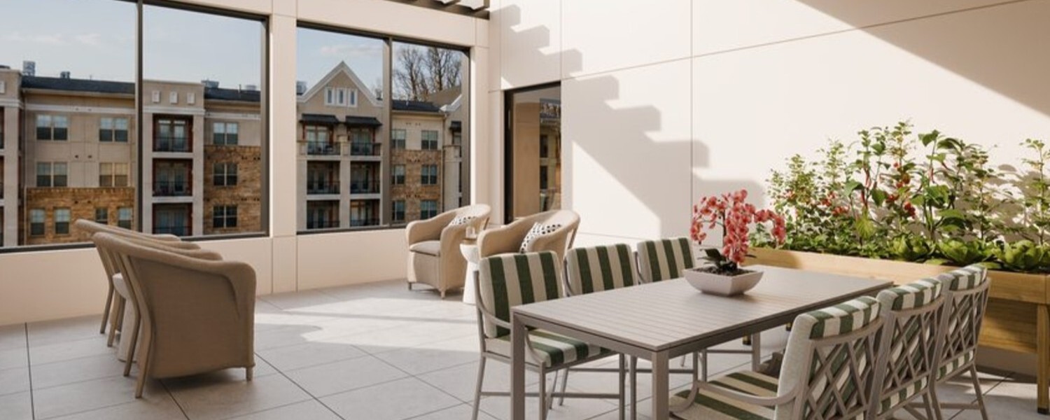 A sunlit outdoor terrace features cozy seating, a dining table, and blooming plants with city views through large windows, showcasing the resort-style senior amenities in Sandy Springs, GA.