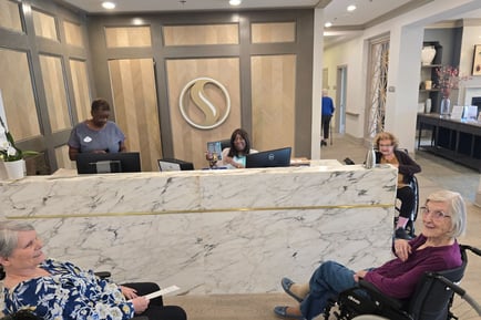 Two staff members work at a marble front desk reception area while two elderly residents in wheelchairs visit nearby, in the lobby of a senior living community.