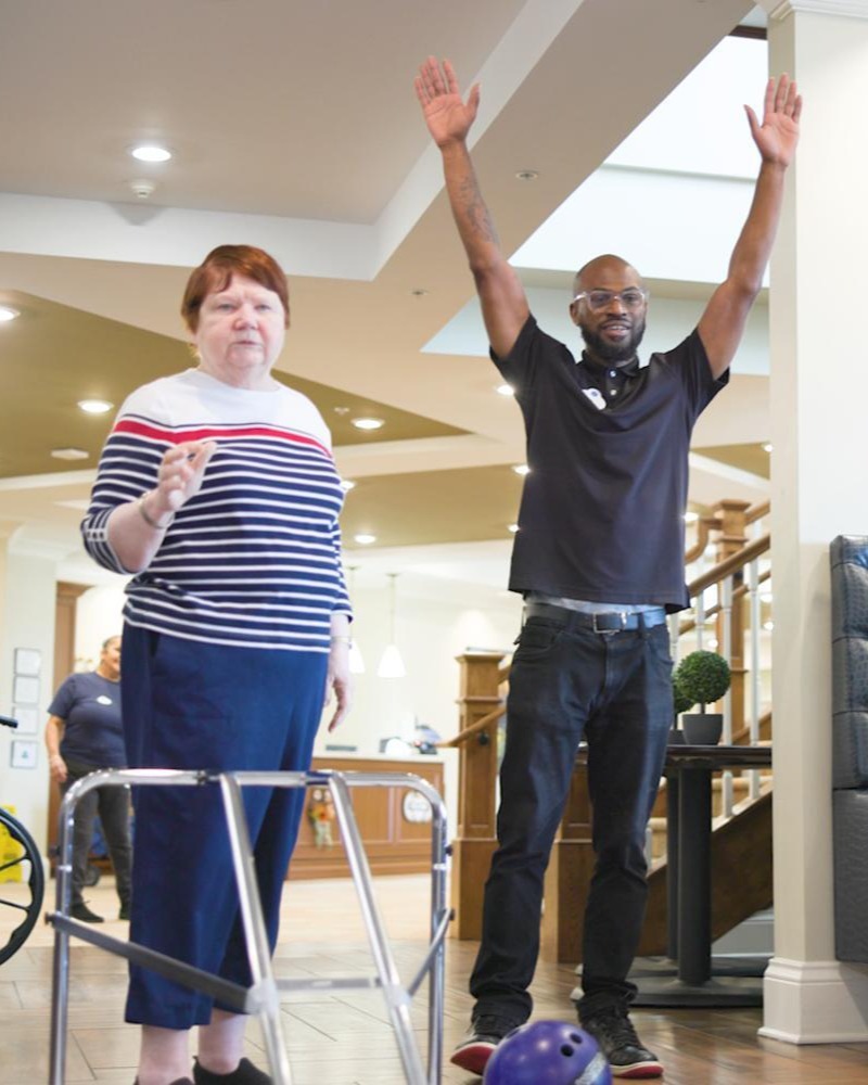A resident and a staff member celebrate a great roll during an indoor bowling activity in the lobby at Arbor Terrace South Forsyth, highlighting the fun, active lifestyle of senior living in Suwanee, GA.