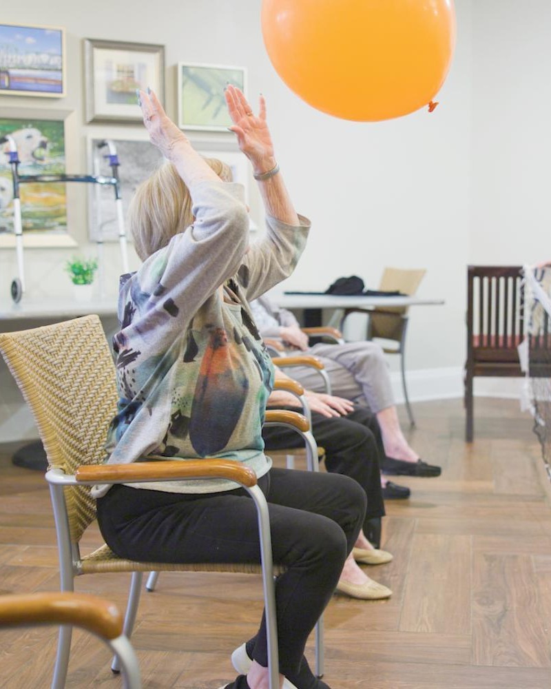 A resident reaches up to tap a large balloon during a seated fitness class at Arbor Terrace South Forsyth, showcasing the wellness-focused, active programming offered at this senior living community in Suwanee, GA.