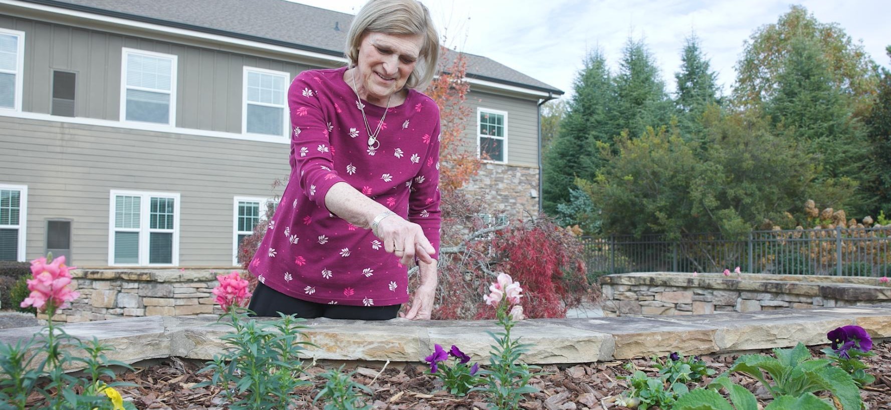 A woman tends to colorful flowers in a stone-edged garden bed outside Arbor Terrace South Forsyth, enjoying the nature-focused outdoor spaces of this senior living community in Suwanee, GA.
