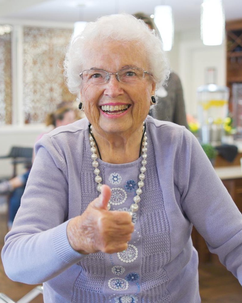 A cheerful older woman gives a thumbs up in a bright common area at Arbor Terrace South Forsyth, capturing the joyful spirit of senior living in Suwanee, GA.