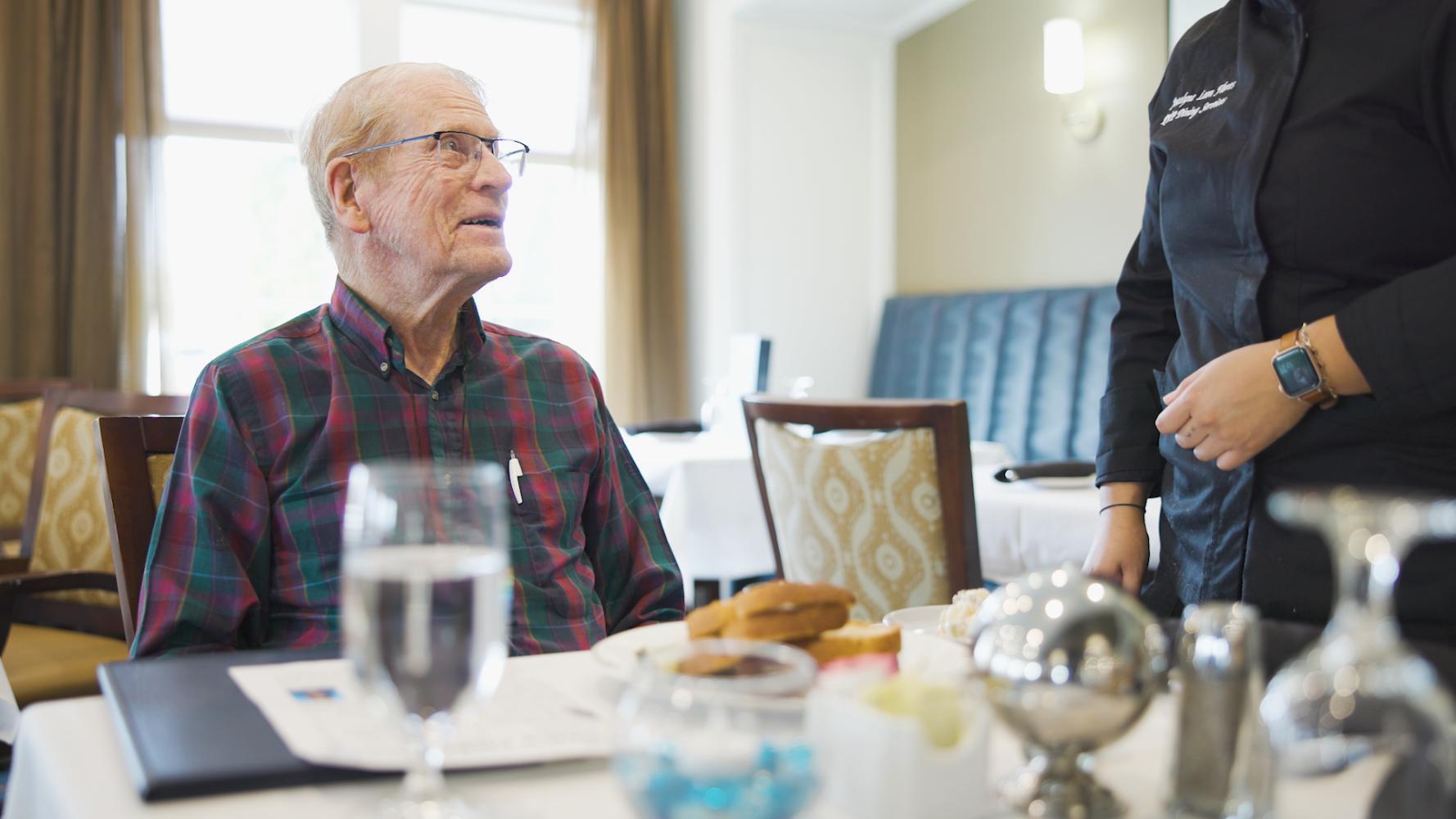 A staff member attends to a resident during a dining service at a warmly set table at Arbor Terrace South Forsyth assisted living community.