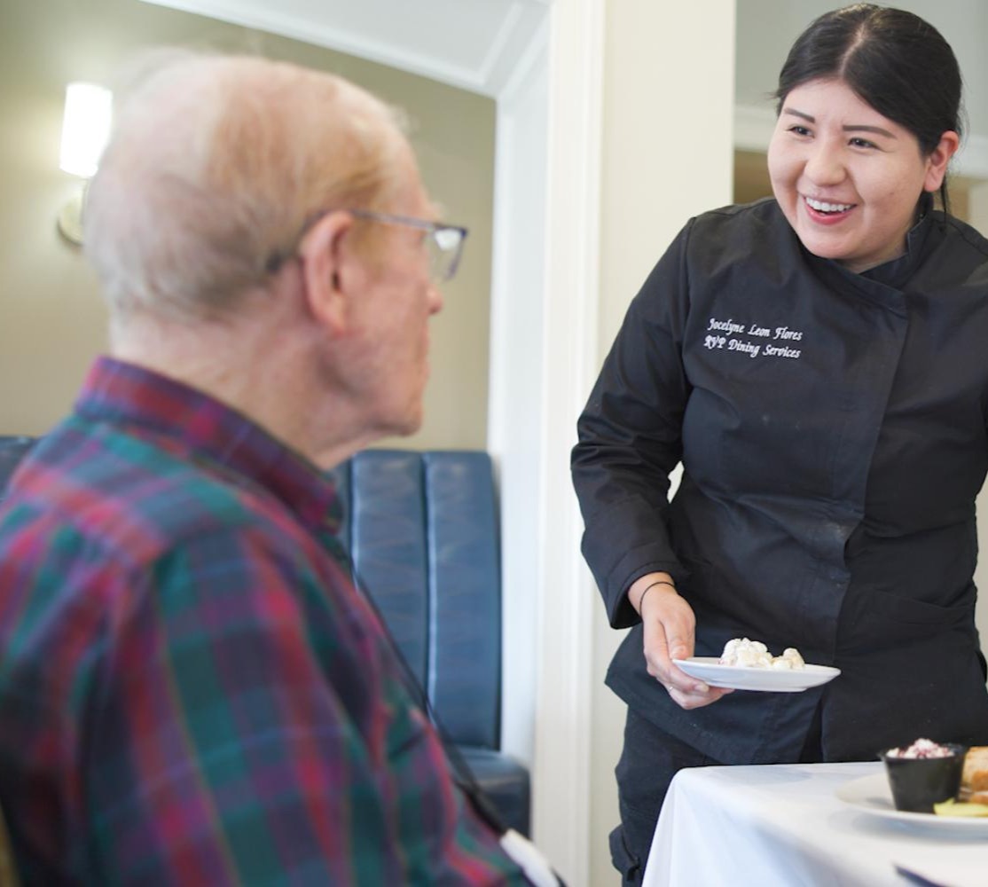A smiling dining staff member serves a plate to a resident in the warm, restaurant-style dining room at Arbor Terrace South Forsyth, highlighting the personalized senior care Suwanee GA families value.