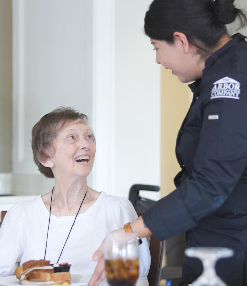 A smiling older woman laughs with an Arbor Company dining staff member during a meal, reflecting the warm, attentive service that defines senior living in Suwanee, GA.