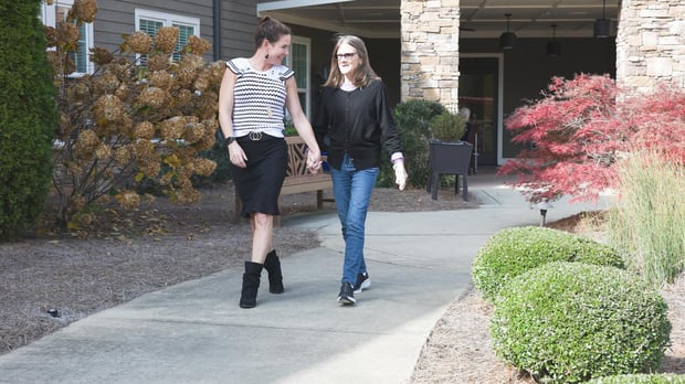Two women walk along a landscaped pathway outside Arbor Terrace South Forsyth, enjoying the welcoming outdoor surroundings of this senior living community in Suwanee, GA.