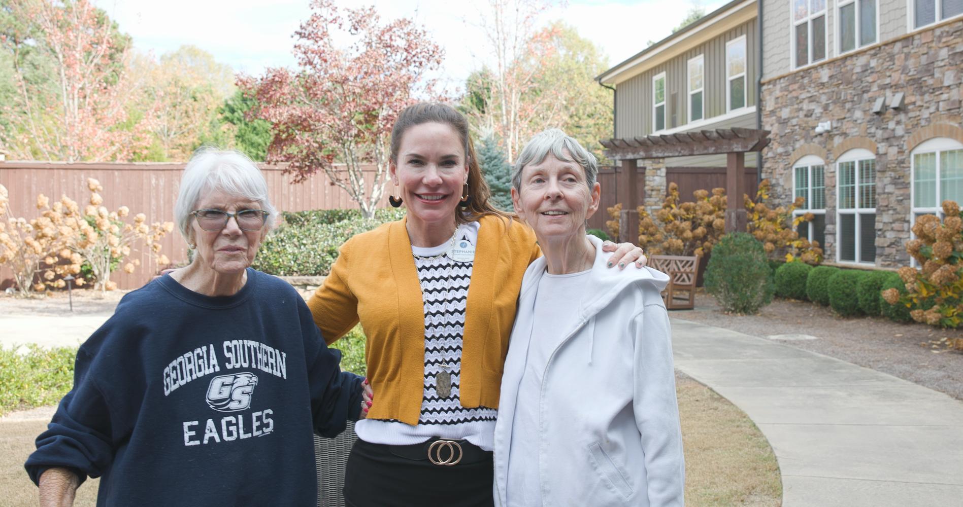 Three people pose together with warm smiles near the stone exterior and autumn foliage at Arbor Terrace South Forsyth.