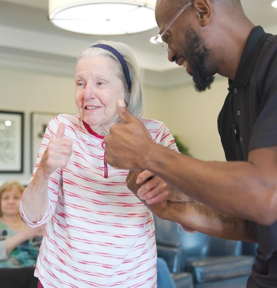 A smiling older woman gives a thumbs up alongside a staff member during an activity at Arbor Terrace South Forsyth, reflecting the warm, engaged spirit of senior living in Suwanee, GA.