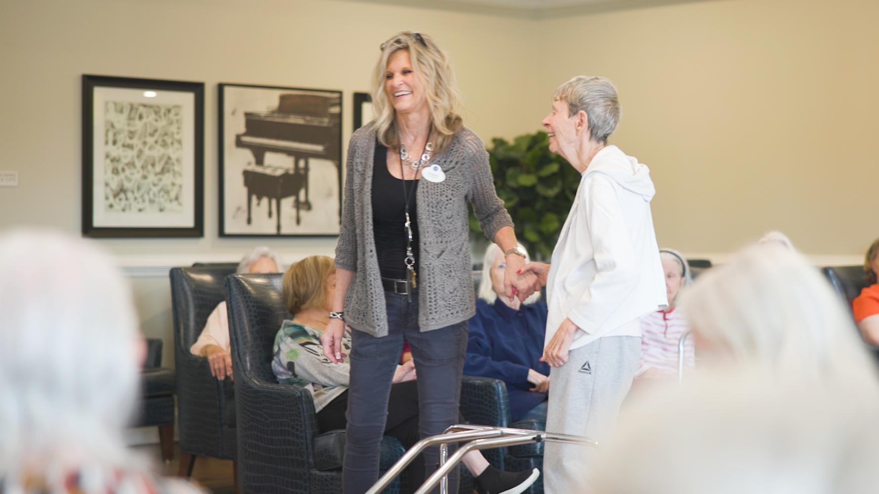 A staff member warmly greets residents gathered in a comfortable lounge area at this assisted living community in Suwanee, GA, with a grand piano print adding a touch of elegance to the space.