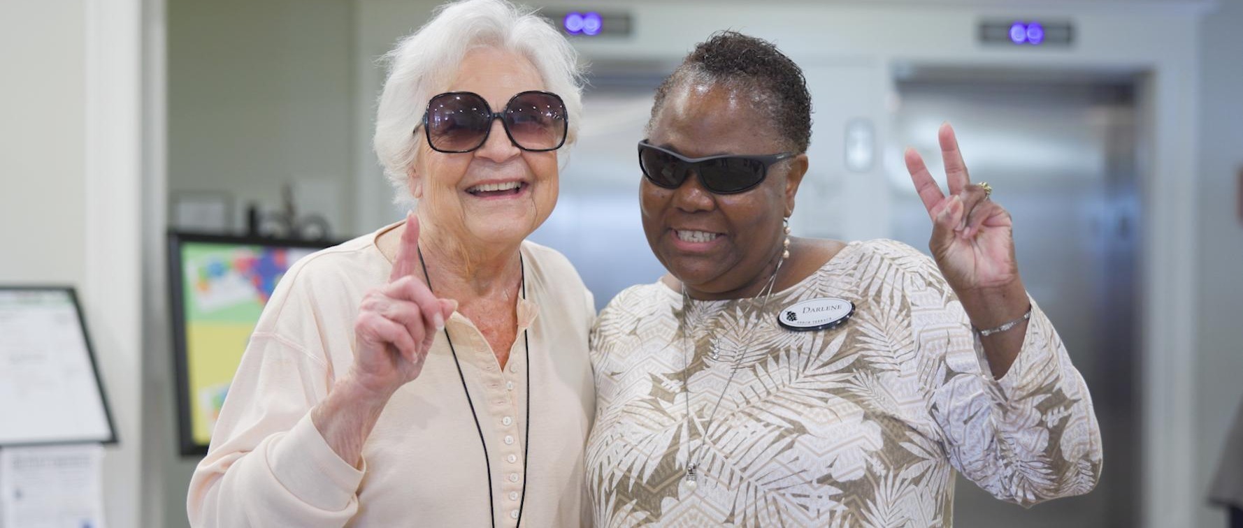 Two women laugh and pose playfully near the elevators at Arbor Terrace South Forsyth, showcasing the joyful, connected lifestyle of senior living in Suwanee, GA.