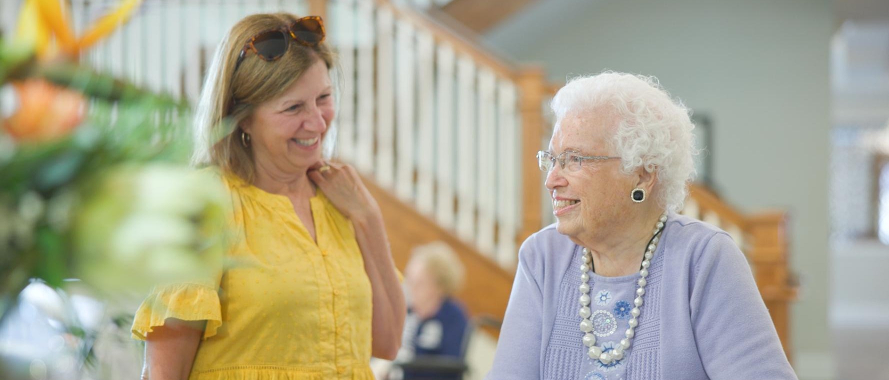Two women share a warm smile in a bright, welcoming common area at Arbor Terrace South Forsyth, a senior living community in Suwanee, GA, with a staircase and lush greenery visible in the background.