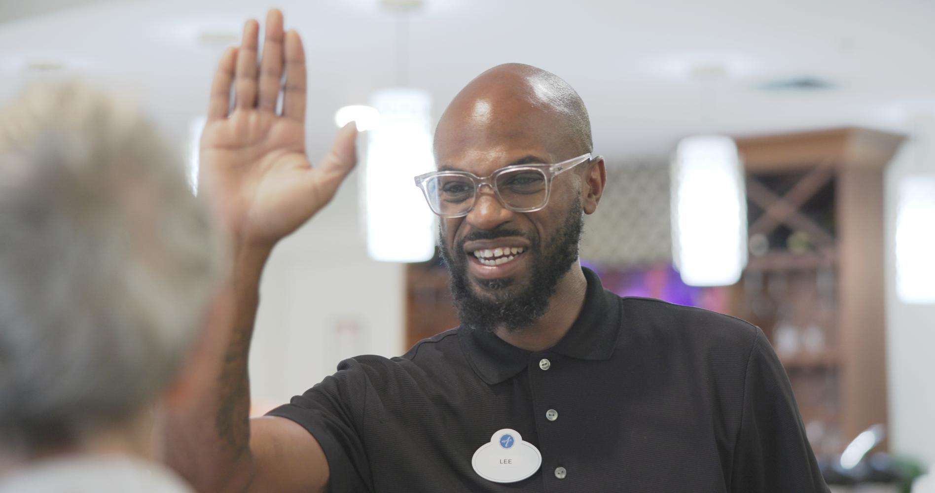 A staff member waves cheerfully in a bright, welcoming common area at Arbor Terrace, a caring assisted living community in Suwanee, GA.