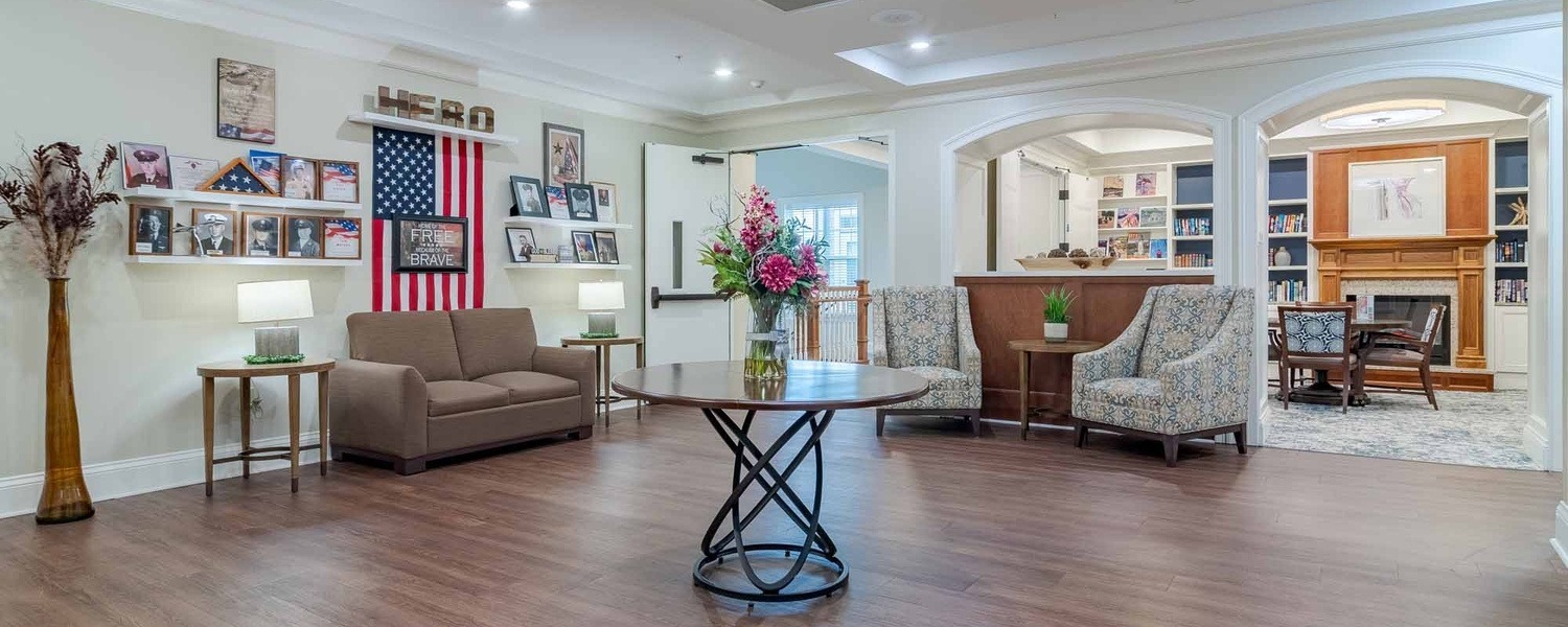 A spacious, welcoming lobby at Arbor Terrace South Forsyth features hardwood floors, comfortable seating, fresh flowers, and classic decor, setting a warm tone for senior living in Suwanee, GA.
