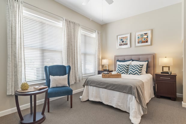 A bright, spacious bedroom apartment at Arbor Terrace South Forsyth features large windows, neutral tones, and tasteful furnishings, offering comfortable senior living in Suwanee, GA.