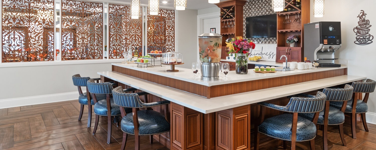 A stylish bistro-style cafe and bar area at Arbor Terrace South Forsyth features warm wood cabinetry, blue upholstered seating, and decorative screens, offering a welcoming gathering spot for senior living in Suwanee, GA.
