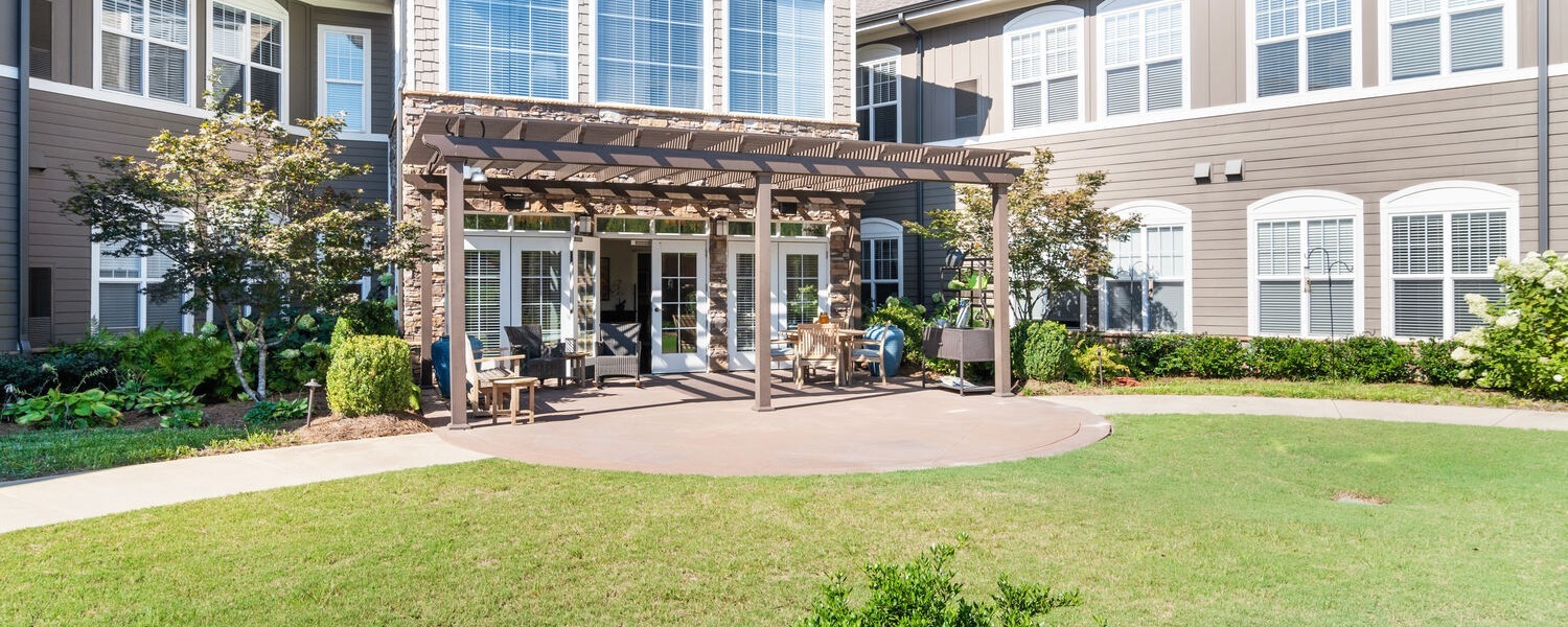 A peaceful outdoor courtyard with a pergola, patio seating, and lush green lawns at Arbor Terrace, a nature-focused senior living community in Suwanee, GA.