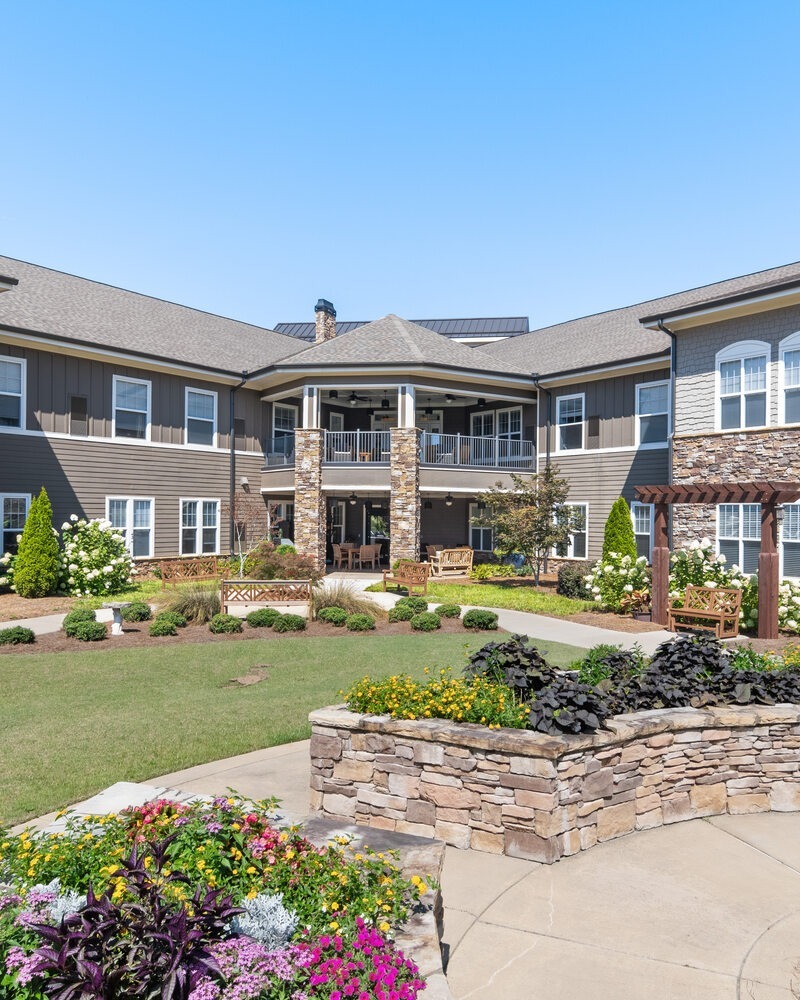 The beautifully landscaped courtyard at Arbor Terrace South Forsyth features colorful gardens, stone planters, a covered patio, and a pergola, creating an inviting outdoor space for senior living in Suwanee, GA.