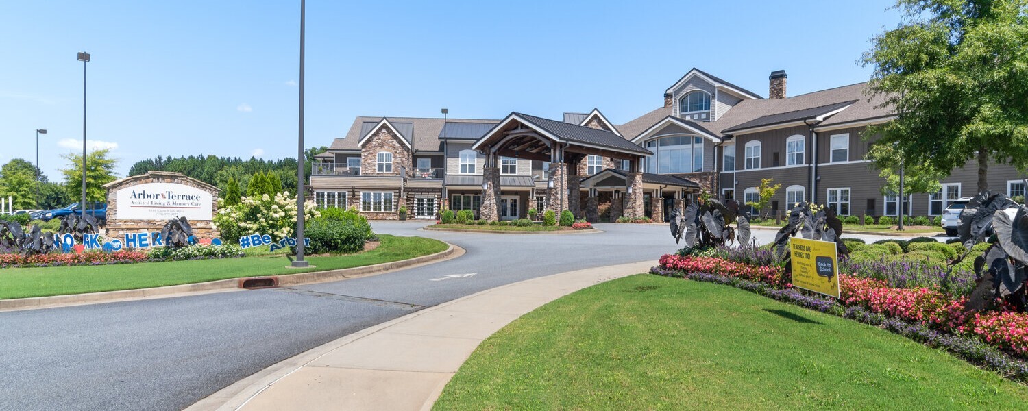 The welcoming entrance of Arbor Terrace South Forsyth features lush landscaping, colorful blooms, and a grand stone-accented building, making it a standout senior living community in Suwanee, GA.