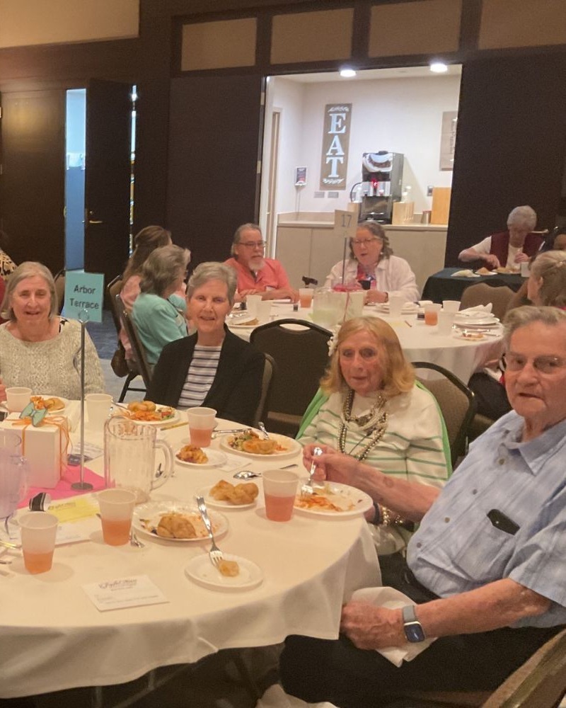 A group of residents and a staff member share smiles and a meal together at a community dining event at Arbor Terrace South Forsyth, enjoying the social spirit of senior living in Suwanee, GA.