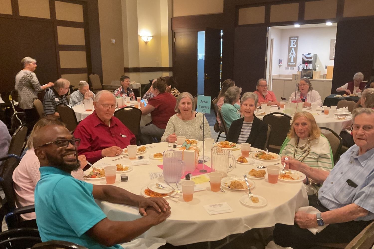 A group of residents and a staff member share smiles and a meal together at a community dining event at Arbor Terrace South Forsyth, enjoying the social spirit of senior living in Suwanee, GA.
