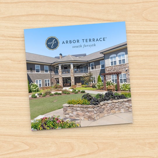 Square photo of Arbor Terrace South Forsyth stone building exterior with landscaped garden wall and manicured shrubs, displayed on a printed card against a wood-grain background.