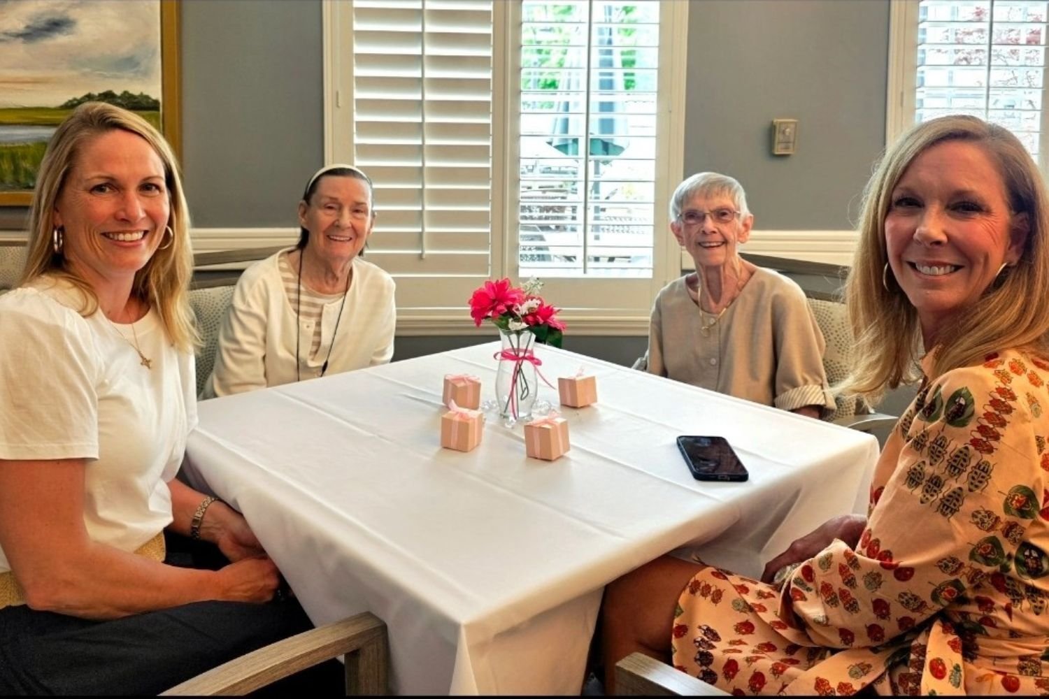residents and family members smile and sit around a table. the table has a vase with pink flowers in it and 4 little gift boxes wrapped in baby pink paper
