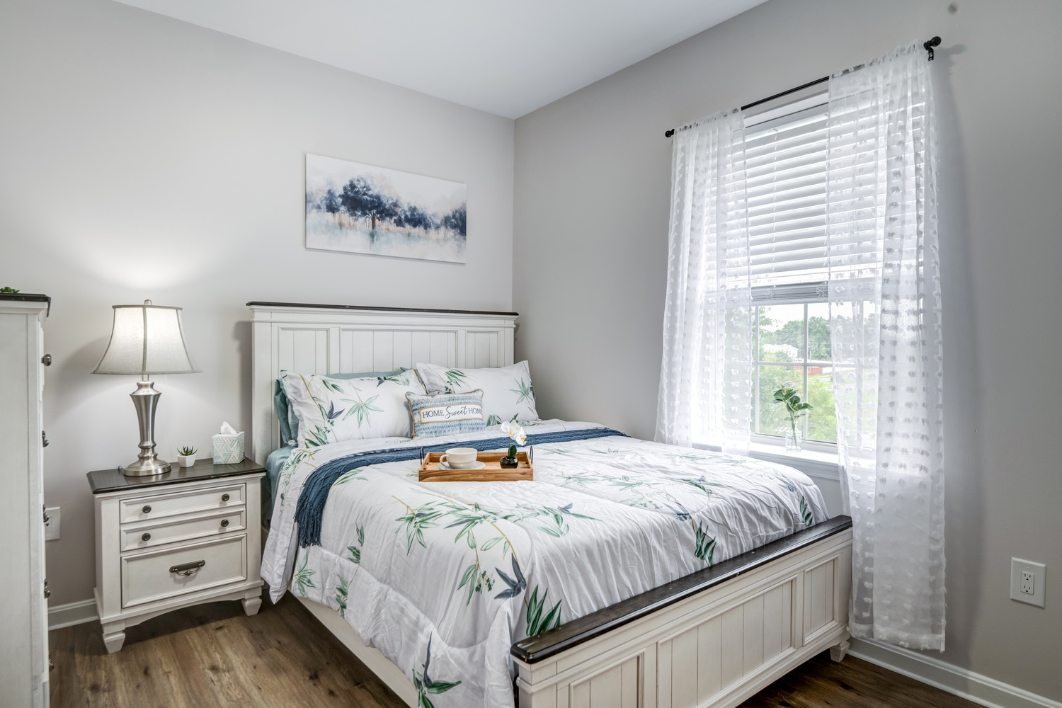 A bedroom in an apartment at Arbor Terrace Sudley Manor with a white bed frame, leafy print bedding, a breakfast tray, a nightstand with a lamp, and sheer curtains at a sunny window, showcasing an apartment at this senior living community in Manassas, VA.