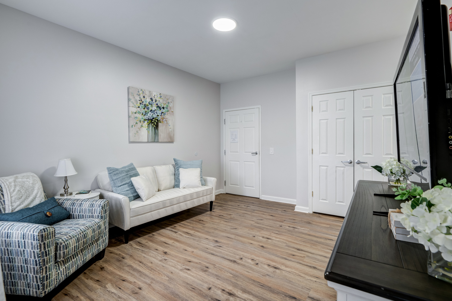 The living area of an apartment at Arbor Terrace Sudley Manor with a cream sofa, patterned armchair, floral artwork, and wide plank flooring, showcasing an apartment at this senior living community in Manassas, VA.