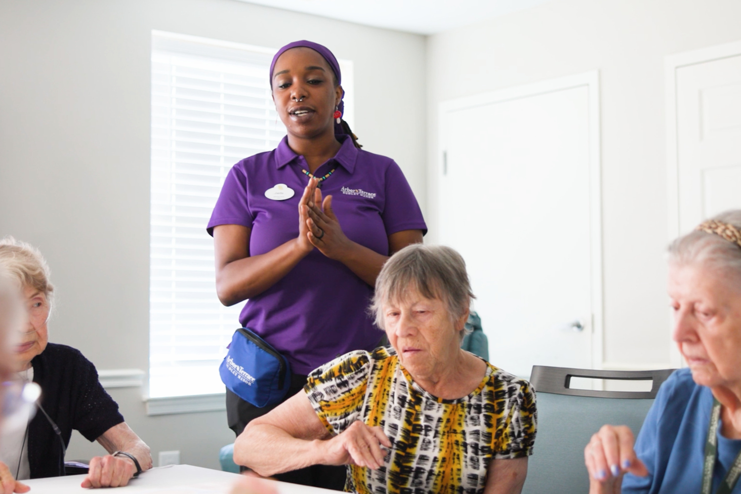 A team member in a purple polo talks with three residents seated around a table during a group activity at Arbor Terrace Sudley Manor, offering friendly senior care in Manassas, Virginia, and senior activities in Northern Virginia.