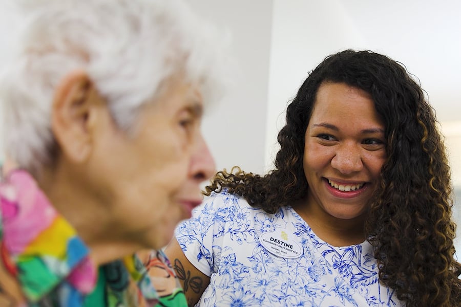 A smiling woman with long curly hair in a blue and white floral top looks toward a woman with white hair in a colorful print top at Arbor Terrace Sudley Manor. Warm connections are part of senior living in Manassas, VA.