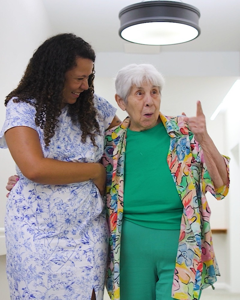 A laughing woman with curly hair links arms with an animated older woman in a colorful floral jacket as they share a lively conversation, a joyful moment that reflects senior living Haymarket VA and the top senior living communities Manassas has to offer.