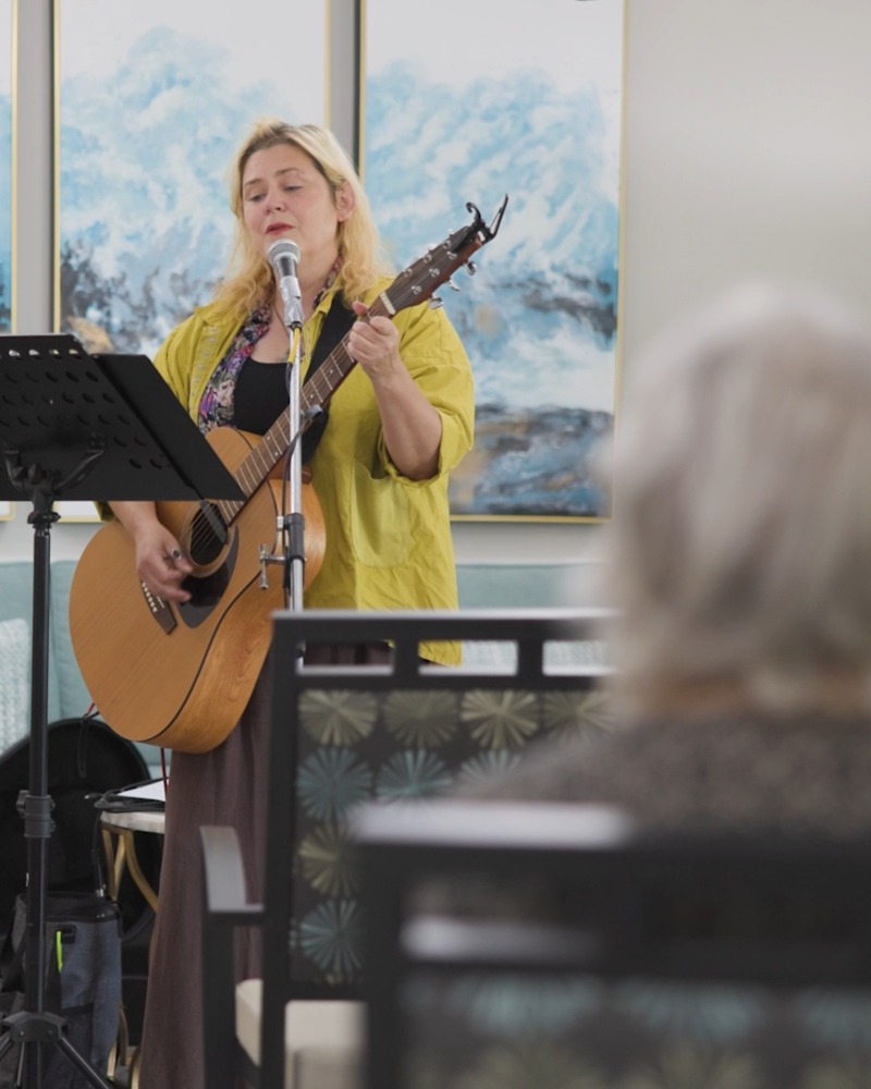 A woman in a mustard jacket sings and plays acoustic guitar at a microphone in a bright room with coastal artwork, a lively scene of senior activities Northern Virginia and the vibrant spirit of senior living near Fairfax VA.