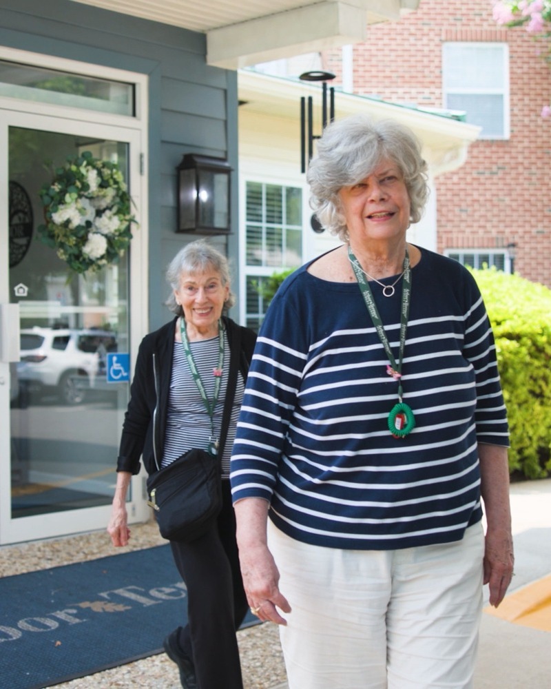 Two smiling older women stroll out of a welcoming front entrance framed by a white floral wreath, brick accents, and lush greenery, a cheerful scene of 55+ senior living Northern Virginia and senior living Manassas VA.