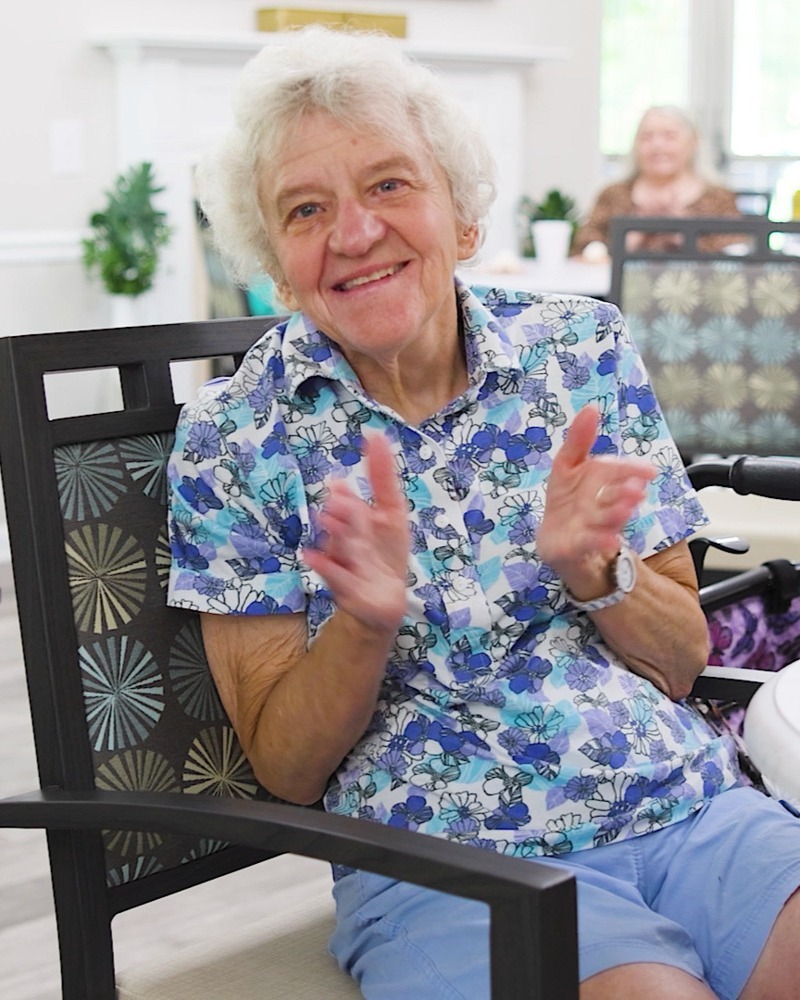 A cheerful older woman in a blue floral blouse claps with delight from her chair in a bright, airy room, sharing the fun of senior activities Northern Virginia and the friendly energy of senior living near Bull Run Manassas.