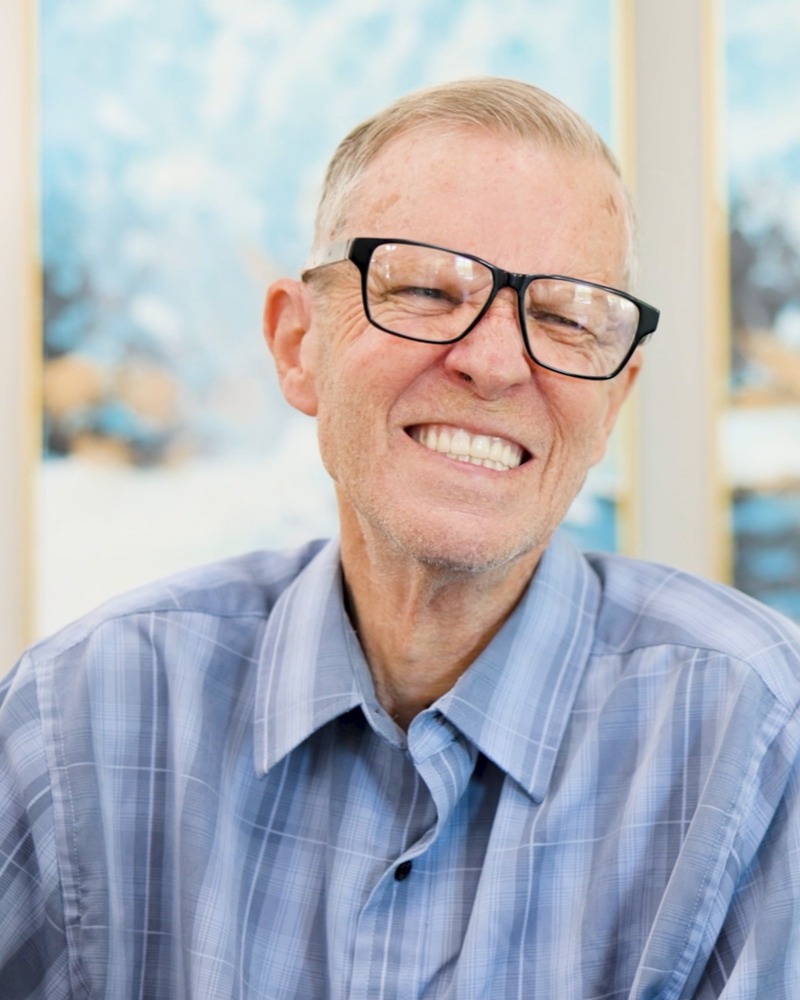 A cheerful older man with glasses and a blue plaid shirt flashes a wide, joyful grin in front of soft blue artwork, a bright moment that reflects the top senior living communities Manassas has to offer and senior living near Fairfax VA.