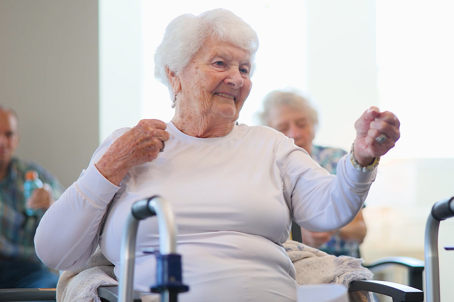 A woman with white hair and grins while keeping the beat in a seated exercise class at Arbor Terrace Sudley Manor. Staying active is part of wellness-focused senior living and an active senior lifestyle in Manassas, VA.